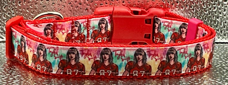 May include: Red dog collar featuring a repeating pattern of a person in a red football jersey with the number 87. The collar has a red plastic buckle and a metal D-ring.