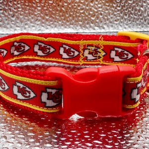 May include: Red dog collar with a repeating pattern of white and red arrowhead logos with a yellow border. The collar has a red plastic buckle and a silver metal D-ring. The collar is designed for a pet.