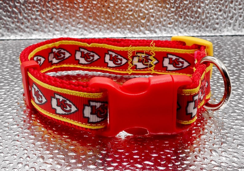 May include: Red dog collar with a repeating pattern of white and red arrowhead logos with a yellow border. The collar has a red plastic buckle and a silver metal D-ring. The collar is designed for a pet.
