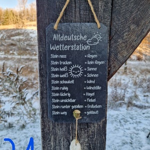 May include: A black slate sign with white text that reads "Altdeutsche Wetterstation" and lists weather conditions based on the state of a stone. The sign is hanging from a wooden post with a string.