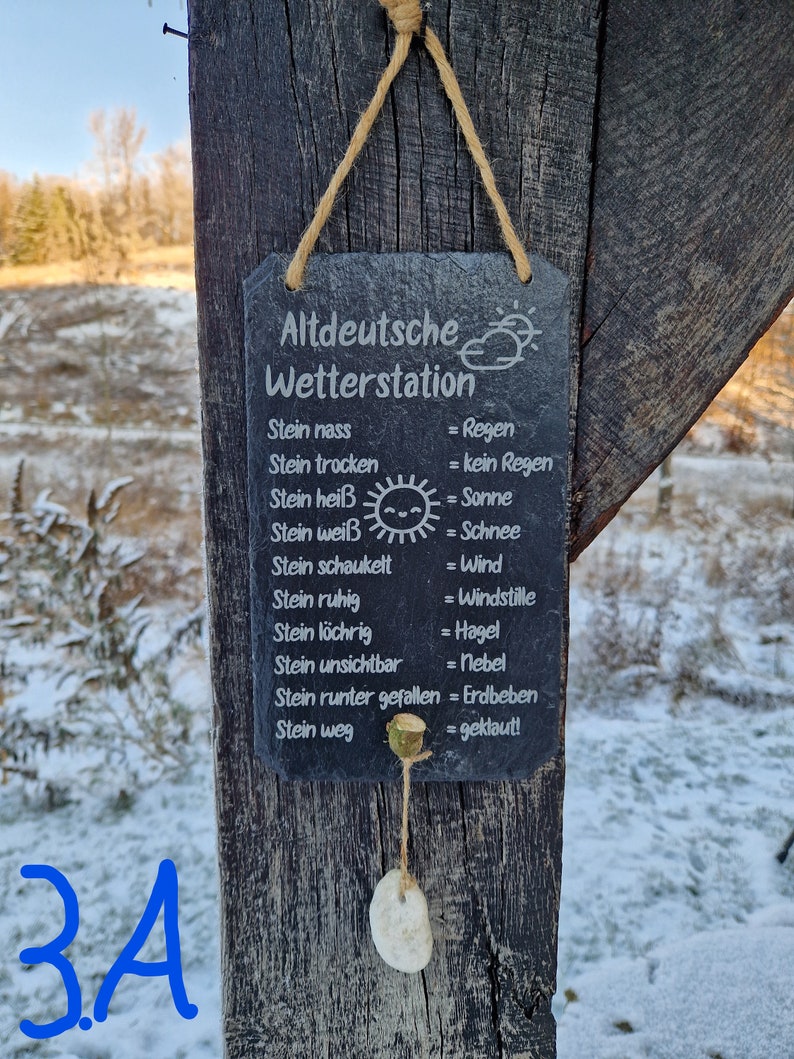 May include: A black slate sign with white text that reads "Altdeutsche Wetterstation" and lists weather conditions based on the state of a stone. The sign is hanging from a wooden post with a string.