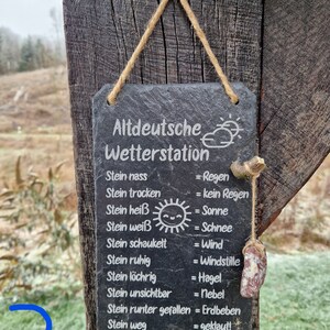 May include: A black slate sign with white text hangs from a wooden post. The sign reads "Altdeutsche Wetterstation" and lists weather conditions in German, such as "Stein nass = Regen" (Stone wet = Rain).