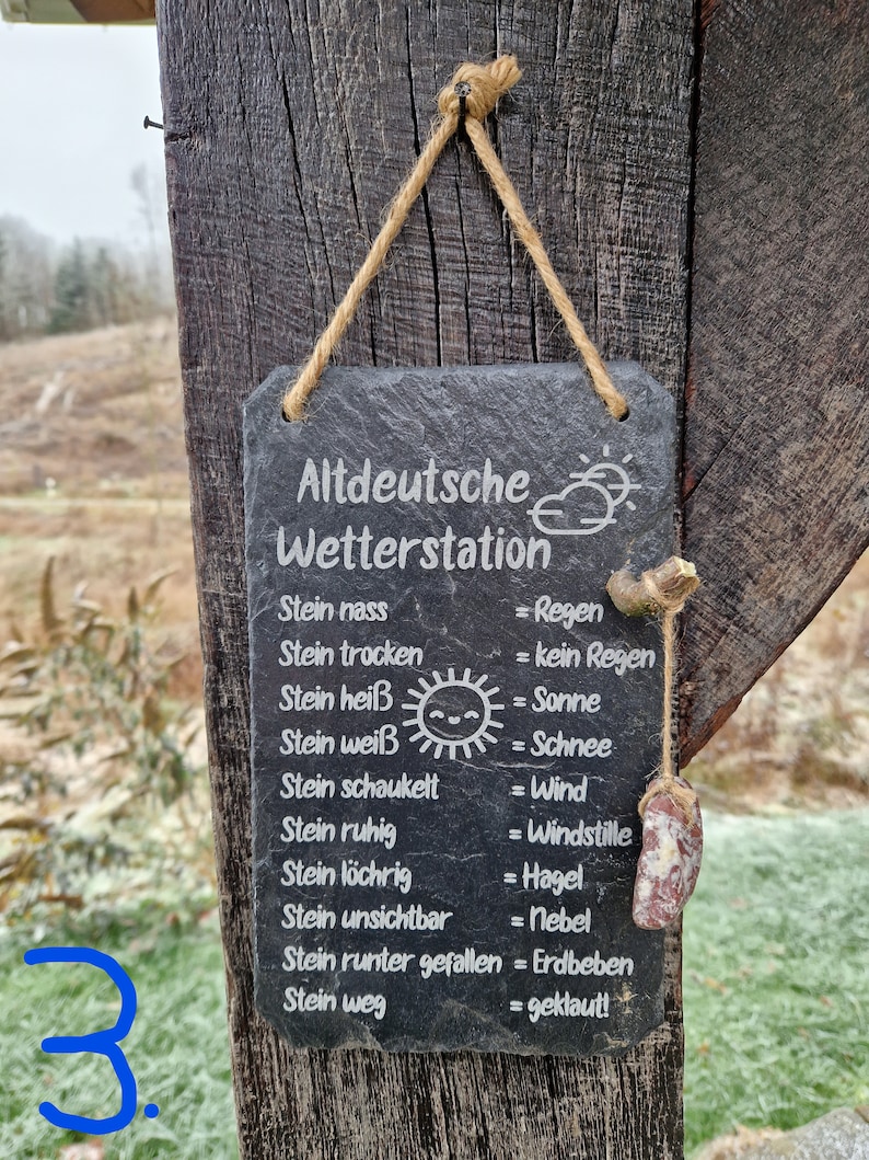May include: A black slate sign with white text hangs from a wooden post. The sign reads "Altdeutsche Wetterstation" and lists weather conditions in German, such as "Stein nass = Regen" (Stone wet = Rain).