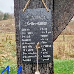 May include: A black slate sign with white text that reads "Altdeutsche Wetterstation" and lists weather conditions based on the state of a stone. The sign is hanging from a wooden post with a rope.