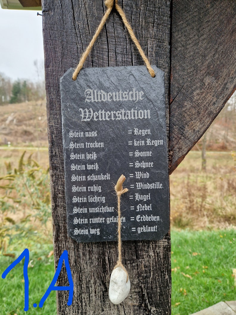 May include: A black slate sign with white text that reads "Altdeutsche Wetterstation" and lists weather conditions based on the state of a stone. The sign is hanging from a wooden post with a rope.