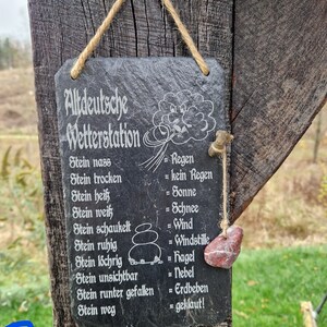 May include: A black slate sign with white text that reads "Altdeutsche Wetterstation" and lists various weather conditions in German with their corresponding meanings in English. The sign is hanging from a wooden post with a piece of twine. A small red stone is hanging from the twine.
