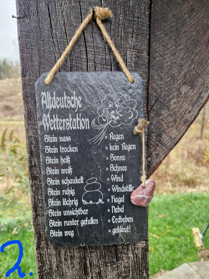 May include: A black slate sign with white text that reads "Altdeutsche Wetterstation" and lists various weather conditions in German with their corresponding meanings in English. The sign is hanging from a wooden post with a piece of twine. A small red stone is hanging from the twine.