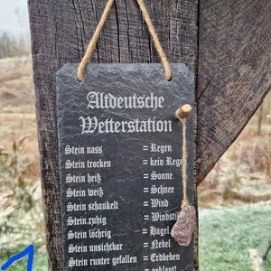 May include: A black slate sign with white text that reads "Altdeutsche Wetterstation" and lists weather conditions based on the state of a stone. The sign is hanging on a wooden post with a brown rope.