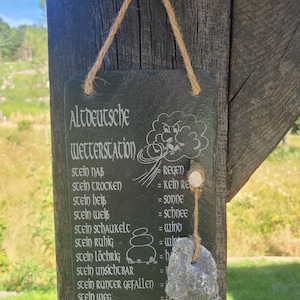 May include: A black slate sign with white text that reads "Altdeutsche Wetterstation" and lists different weather conditions in German. The sign is hanging from a wooden beam with a rope. A small gray rock is hanging from the bottom of the sign.