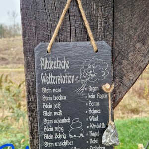 May include: A black slate sign with white text that reads "Altdeutsche Wetterstation" and lists weather conditions based on the state of a stone. The sign is hanging from a wooden post with twine.