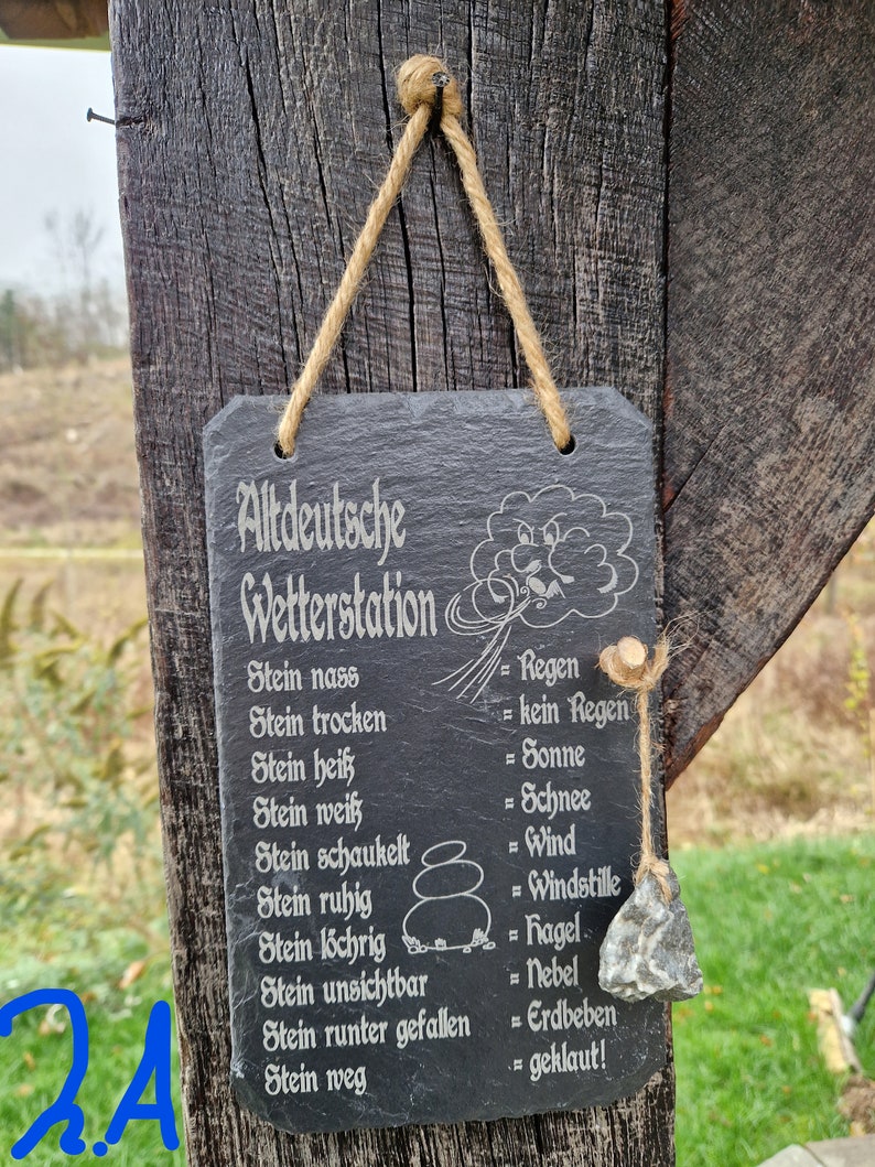 May include: A black slate sign with white text that reads "Altdeutsche Wetterstation" and lists weather conditions based on the state of a stone. The sign is hanging from a wooden post with twine.