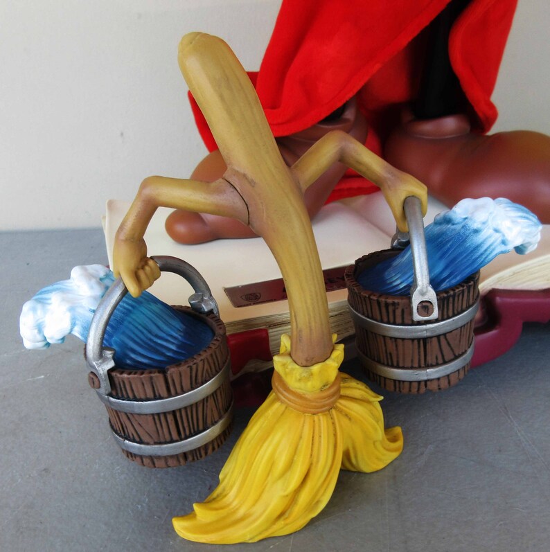 May include: A wooden broom with a yellow bristle brush and two brown buckets with blue water and white foam. The broom is attached to a wooden figure with a red robe.