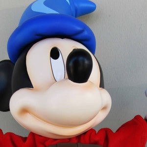 May include: A blue and white sorcerer's hat sits on top of a Mickey Mouse character's head. The character is wearing a red robe and has a big smile. The character is holding out a white gloved hand.