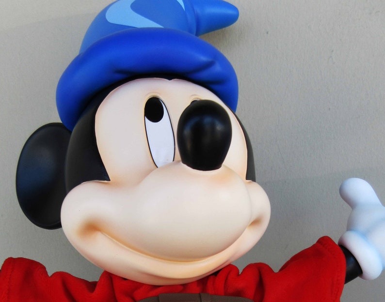 May include: A blue and white sorcerer's hat sits on top of a Mickey Mouse character's head. The character is wearing a red robe and has a big smile. The character is holding out a white gloved hand.