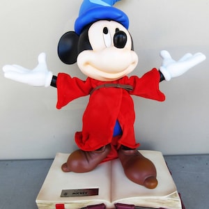 May include: A red and blue sorcerer Mickey Mouse figurine standing on a large open book with the text "Fantasia Mickey" on the front cover.