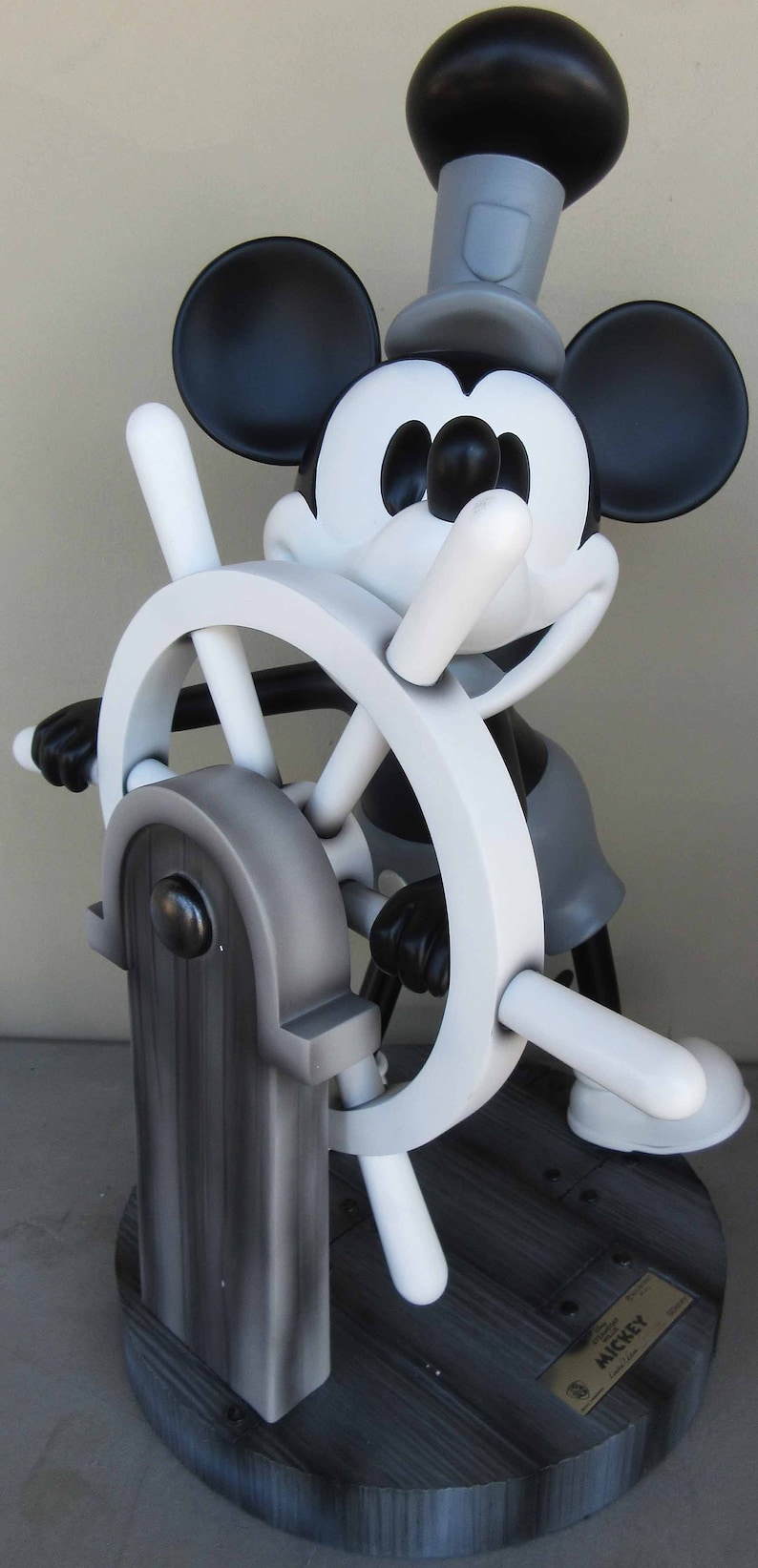 May include: Black and white figurine of Mickey Mouse dressed as a sailor, holding a white ship's wheel with a black base. The base has a plaque that reads "Mickey".