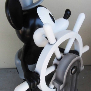 May include: A black and white figurine of Mickey Mouse dressed as a sailor, holding a white ship's wheel. The figurine is standing on a black wooden base.