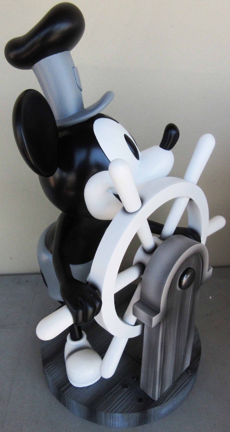 May include: A black and white figurine of Mickey Mouse dressed as a sailor, holding a white ship's wheel. The figurine is standing on a black wooden base.