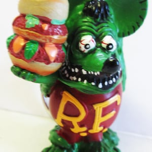 May include: Green monster figure with a toothy grin, holding a triple-decker burger. The figure is wearing a red shirt with the letters "RF" in yellow.