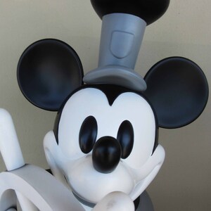 May include: A black and white Mickey Mouse figurine wearing a black top hat with a gray band. The figurine is holding a white wooden steering wheel.