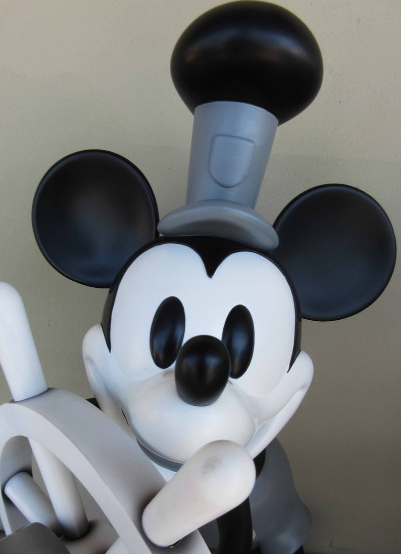 May include: A black and white Mickey Mouse figurine wearing a black top hat with a gray band. The figurine is holding a white wooden steering wheel.