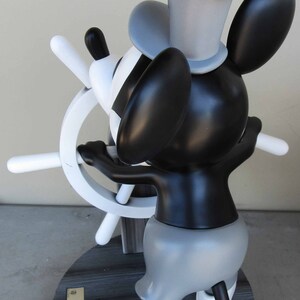 May include: A black and white Mickey Mouse figurine holding a ship's wheel. The figurine is wearing a captain's hat and has a gray and black body. The figurine is standing on a wooden base with the words "The Original Mickey Mouse" and "Mickey" printed on a gold plaque.
