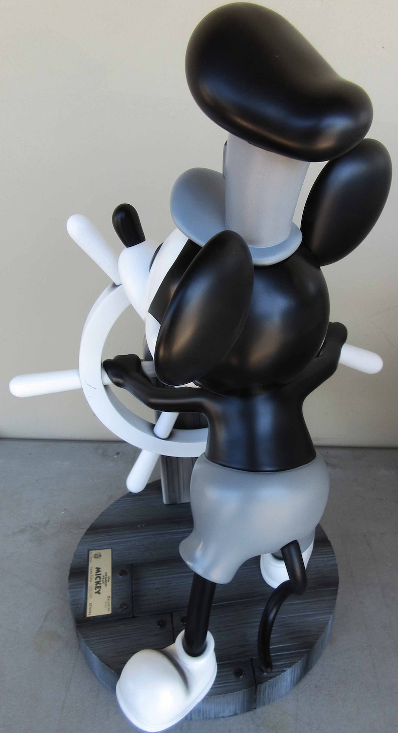 May include: A black and white Mickey Mouse figurine holding a ship's wheel. The figurine is wearing a captain's hat and has a gray and black body. The figurine is standing on a wooden base with the words "The Original Mickey Mouse" and "Mickey" printed on a gold plaque.
