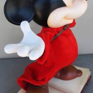 May include: A red and blue sorcerer Mickey Mouse figurine standing on a closed book with the text "Fantasia Mickey" on the cover. The figurine is wearing a blue hat with a white star and a red robe.