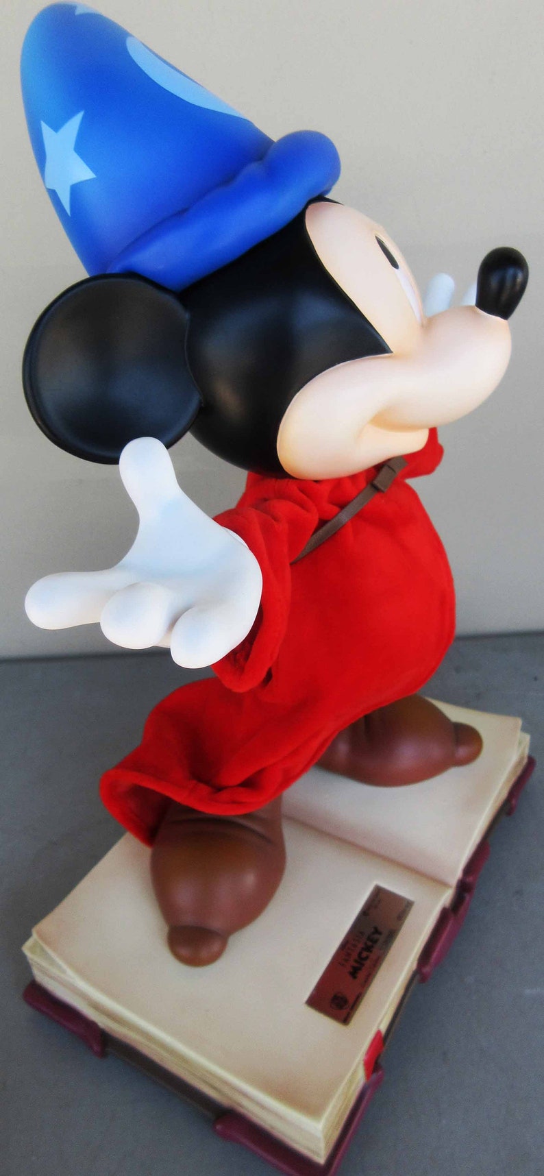 May include: A red and blue sorcerer Mickey Mouse figurine standing on a closed book with the text "Fantasia Mickey" on the cover. The figurine is wearing a blue hat with a white star and a red robe.