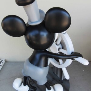 May include: A black and white Mickey Mouse figurine holding a white ship's wheel. Mickey is wearing a black and white sailor hat and a gray shirt. The figurine is standing on a black and white wooden base.