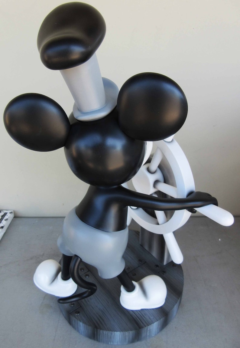 May include: A black and white Mickey Mouse figurine holding a white ship's wheel. Mickey is wearing a black and white sailor hat and a gray shirt. The figurine is standing on a black and white wooden base.