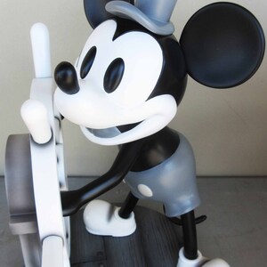 May include: A black and white figurine of Mickey Mouse wearing a gray hat and holding a white steering wheel. The figurine is standing on a wooden base with a plaque that reads "Mickey Mouse".