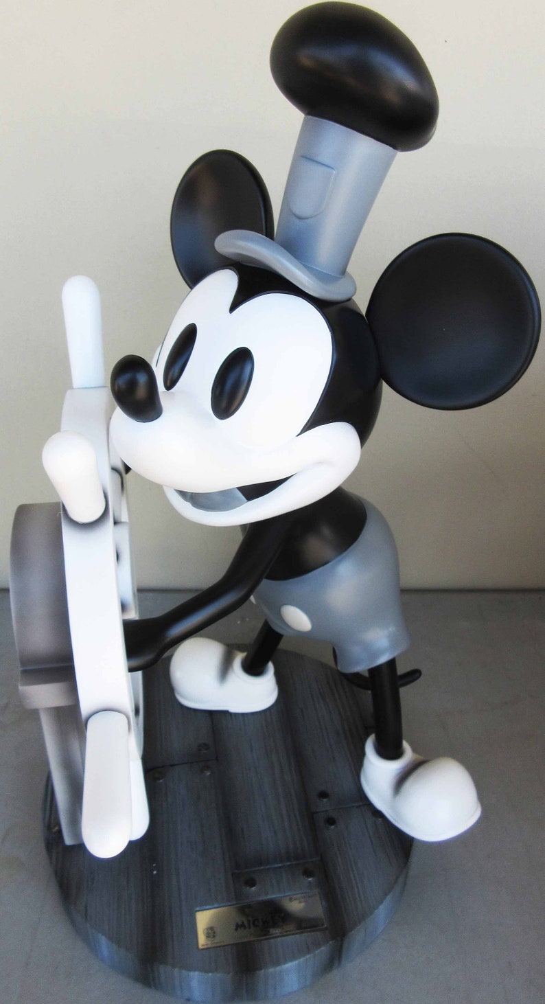 May include: A black and white figurine of Mickey Mouse wearing a gray hat and holding a white steering wheel. The figurine is standing on a wooden base with a plaque that reads "Mickey Mouse".