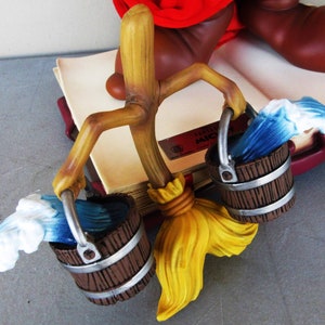 May include: A wooden stick figure holding two brown buckets with blue and white water waves. The stick figure is holding a yellow broom. The figure is sitting on a book with the text "Disney's Mickey Mouse" on the cover.