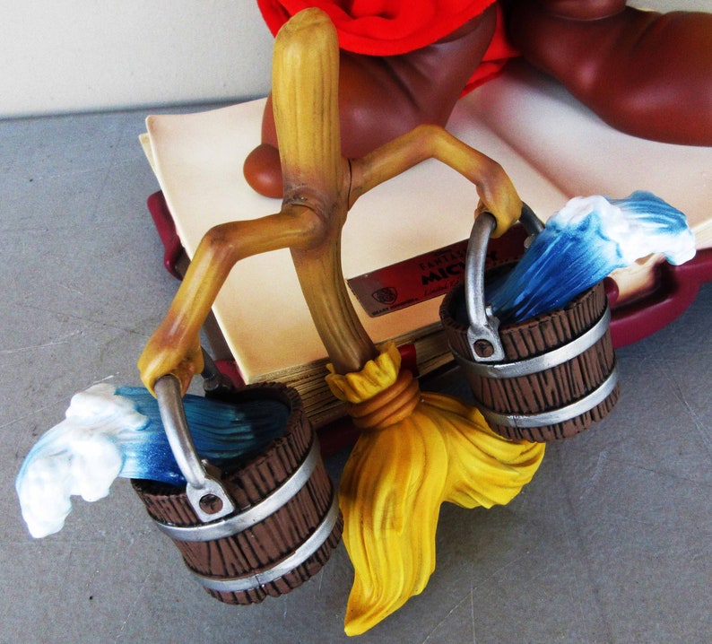 May include: A wooden stick figure holding two brown buckets with blue and white water waves. The stick figure is holding a yellow broom. The figure is sitting on a book with the text "Disney's Mickey Mouse" on the cover.