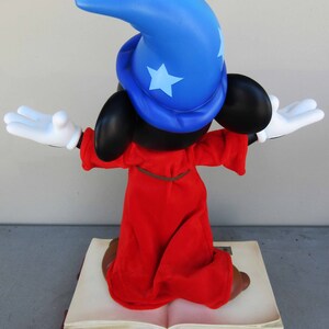 May include: A red and blue sorcerer Mickey Mouse figurine standing on a closed book. Mickey is wearing a blue pointed hat with a white star and a red robe. He is holding his arms out in a magical gesture.