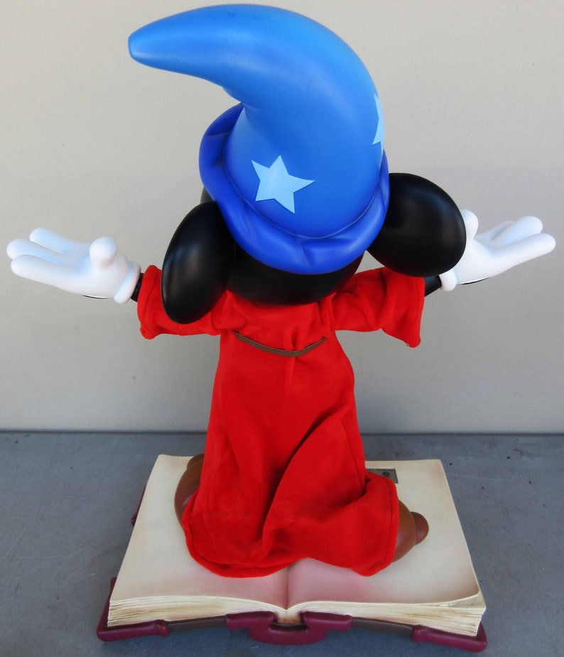 May include: A red and blue sorcerer Mickey Mouse figurine standing on a closed book. Mickey is wearing a blue pointed hat with a white star and a red robe. He is holding his arms out in a magical gesture.