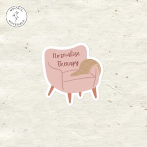 Puede incluir: Una pegatina con una butaca rosa con la frase "Normalize Therapy" impresa. Una manta color beige está sobre la silla. La pegatina tiene un borde blanco y está sobre un fondo texturizado.