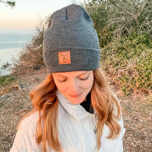 Puede incluir: Un gorro gris con un parche de cuero marrón que presenta un diseño de zorro. El gorro lo lleva una persona con el pelo largo y castaño rojizo.