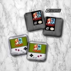 May include: Cornhole game bags featuring a retro video game design. Two gray bags resemble classic game cartridges, while two silver bags are styled like handheld consoles. Each bag displays a pixelated graphic with the text "CORNHOLE".