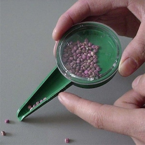May include: A green plastic seed dispenser with a clear lid. The dispenser is filled with small pink and brown seeds. A few seeds have fallen out of the dispenser onto a gray surface.