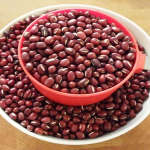 May include: A white bowl filled with red beans, with a red measuring cup filled with red beans on top.
