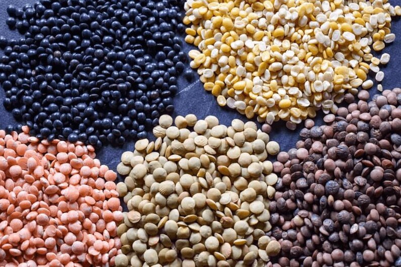 May include: A close-up view of six different types of lentils: black, red, green, brown, yellow, and split. The lentils are arranged in a circle on a dark gray surface.