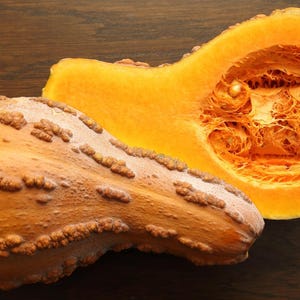 Pode incluir: Uma ab&oacute;bora butternut cortada ao meio, com polpa de cor laranja claro e um exterior marrom e irregular. A ab&oacute;bora est&aacute; sobre uma superf&iacute;cie de madeira marrom escura.