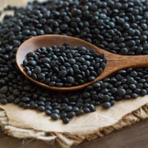 May include: A wooden spoon filled with black lentils, surrounded by more lentils on a brown burlap surface.