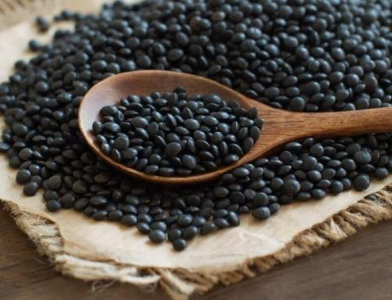 May include: A wooden spoon filled with black lentils, surrounded by more lentils on a brown burlap surface.