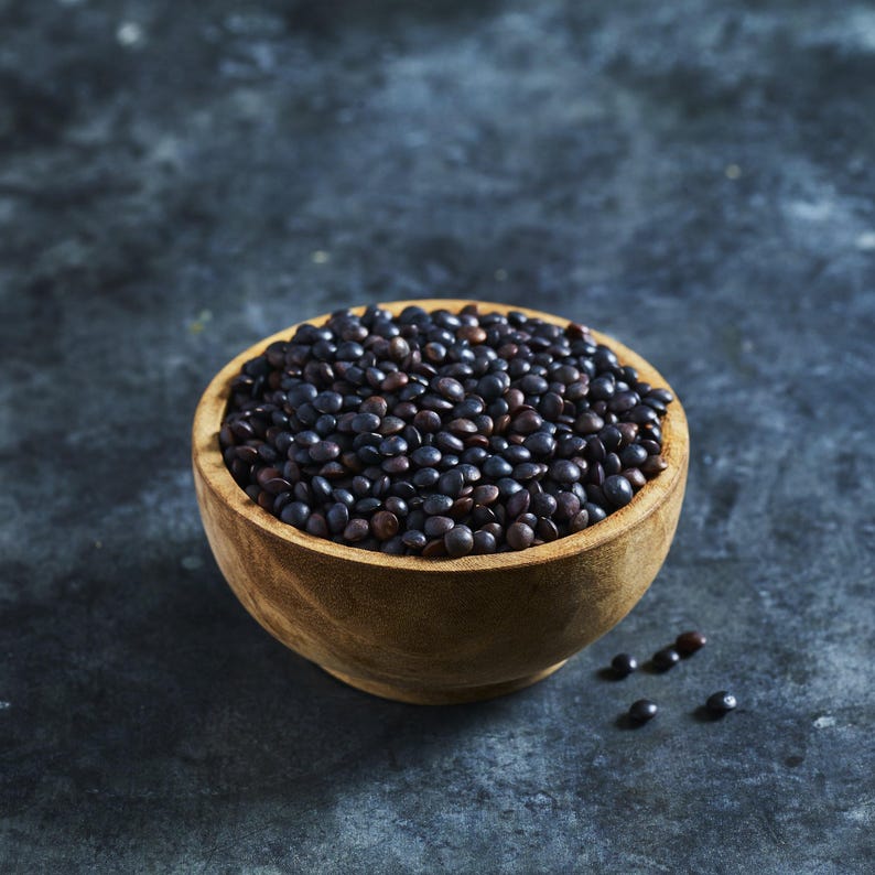 May include: A wooden bowl filled with black lentils. The lentils are a dark, almost black color and are arranged in a pile in the bowl. The bowl is sitting on a gray surface.