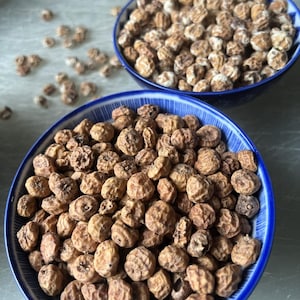 May include: Two blue and white patterned bowls filled with tiger nuts. The tiger nuts are small, round, and have a light brown color. Some nuts are scattered around the bowls. The bowls are on a light gray surface.