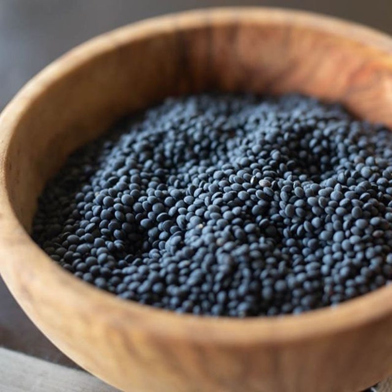 May include: A wooden bowl filled with black lentils. The lentils are a dark grey color and are piled high in the bowl.