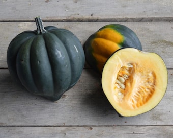 May include: Three acorn squash on a wooden surface. One whole squash is dark green, another is green and yellow, and the third is cut in half, revealing the seeds and orange flesh. The squash are a seasonal food.